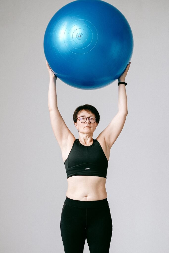 Senior woman in activewear lifting a blue exercise ball high above her head in a studio setting.